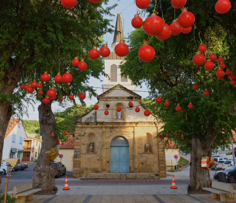 L’image montre une petite église en pierre au centre d’un village européen, entourée d’arbres décorés avec des boules rouges et des cadeaux, ce qui crée une ambiance de Noël. On voit aussi des bancs, des voitures et des maisons autour de la place.
