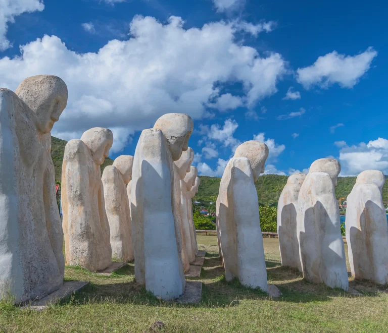 L’image montre des statues blanches penchées face à la mer. Elles représentent des esclaves africains et symbolisent le souvenir et le recueillement. Ce monument se trouve au Mémorial de l’Anse Caffard près du Diamant en Martinique. Il a été créé par Laurent Valère pour rendre hommage aux esclaves morts lors d’un naufrage en 1830.