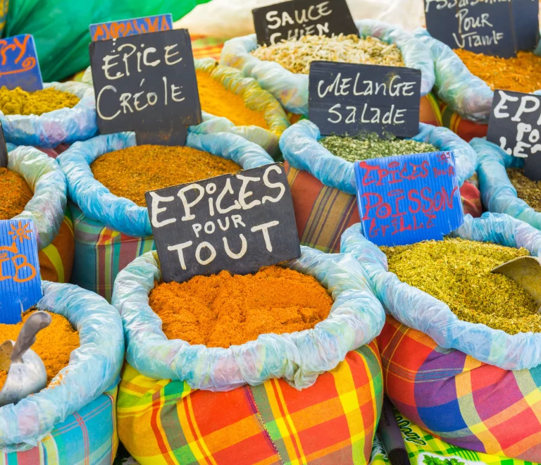 L'image montre des sacs en tissu madras coloré, remplis de divers mélanges d'épices artisanaux, sur un étal de marché traditionnel. On y voit des pancartes écrites à la main en français et en créole, proposant des mélanges spécifiques comme les "Épices Créoles", le "Sel Caraïbe", ou encore la fameuse "Sauce Chien".
