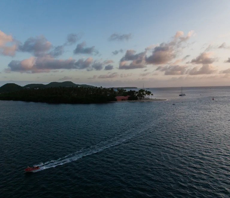L’image montre un petit îlet entouré par la mer avec des palmiers et quelques bateaux. On voit aussi un jet-ski laissant une trace sur l’eau, sous un ciel coloré par le coucher du soleil.